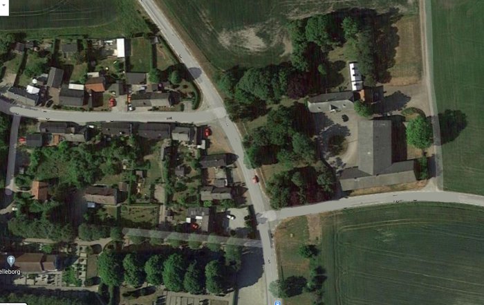Flygbild över en by med en huvudväg som skär genom och anslutande mindre vägar omgivna av byggnader och vegetation.