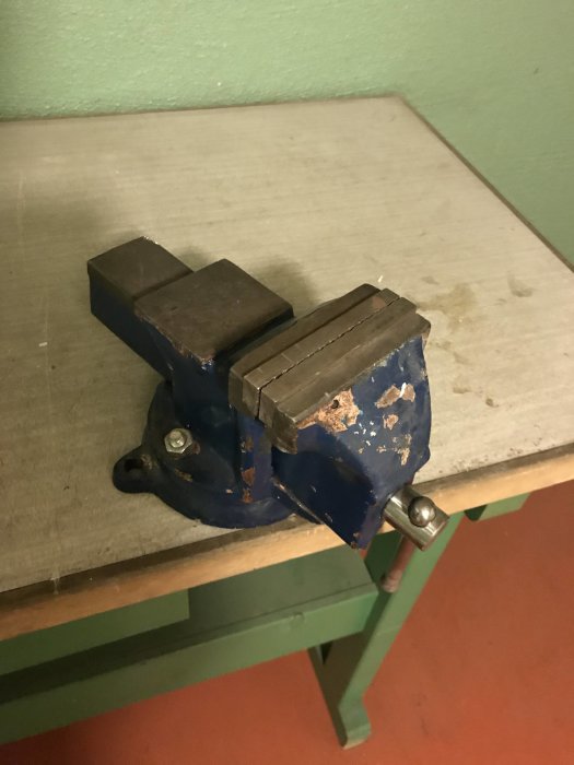 Blått skruvstycke med slitage på arbetsbänk mot grön vägg.