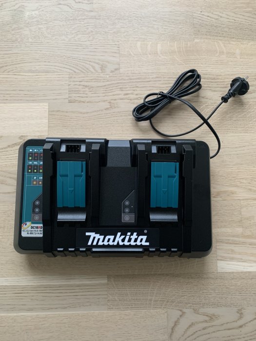 En ny Makita dubbelladdare och batterihållare på ett trägolv, ej inkopplad med strömkabeln bredvid.