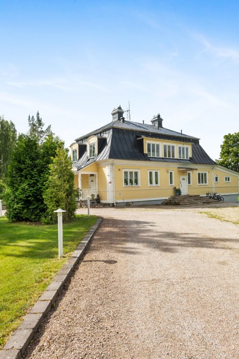 Gult tvåvåningshus från 1905 med svart plåttak och stor grusuppfart framför, omgivet av grönska.