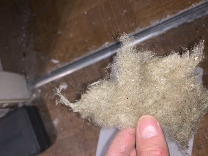 Hand håller en bit äldre isoleringsmaterial mot bakgrund av trägolv och metallspår.