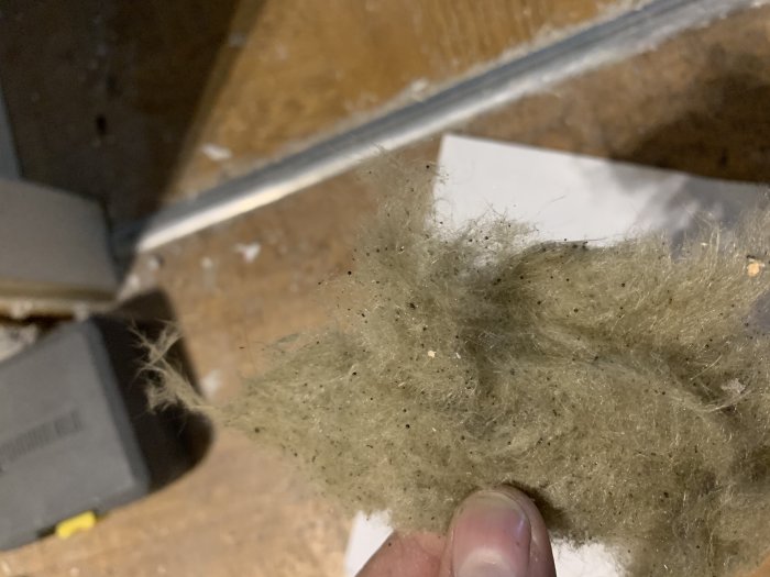 Hand håller gammal isolering, eventuellt stenull eller glasull, på en vind, med osäkerhet om innehåll.