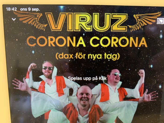 Skärm med bild på tre personer i orangea överdragskläder på svart bakgrund med texten "VIRUZ CORONA (dax för nya tag)