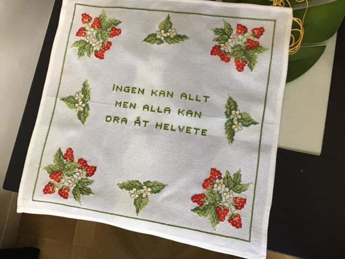 Broderad text "INGEN KAN ALLT MEN ALLA KAN DRÄTT HELVETE" med röda och gröna blommor runtomkring på vit duk.