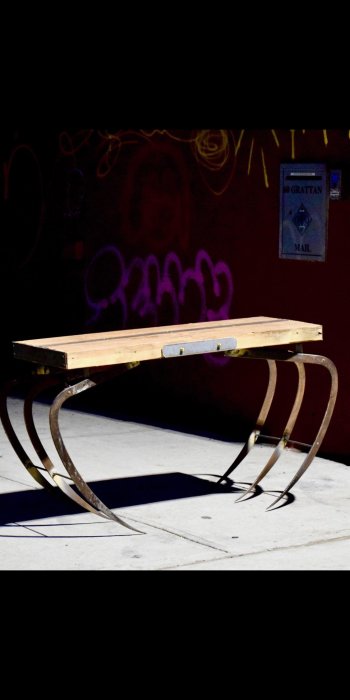 En smal bänk med ett träskiva ovansida och böjda spetsiga metallben mot en vägg med graffiti.