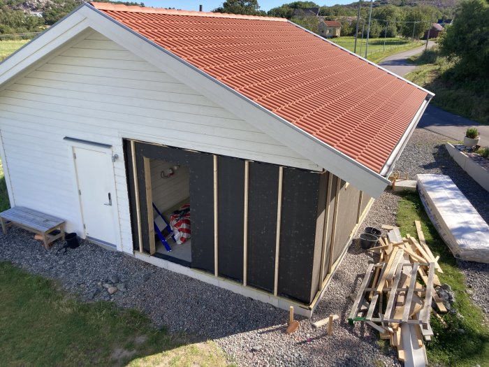 Vit fristående byggnad med asfaboard monterat och reglar för isolering på ena sidan, byggmaterial syns på marken.