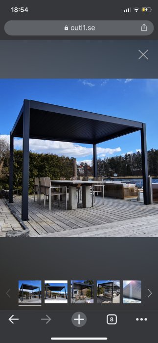 Svart pergola med tak över trädäck vid vatten med utebord och stolar, mot skog och klarblå himmel.