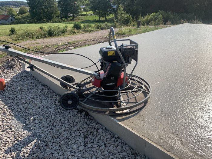 Power trowel på nylagt betonggolv utomhus, med fält av grus och grönska i bakgrunden.