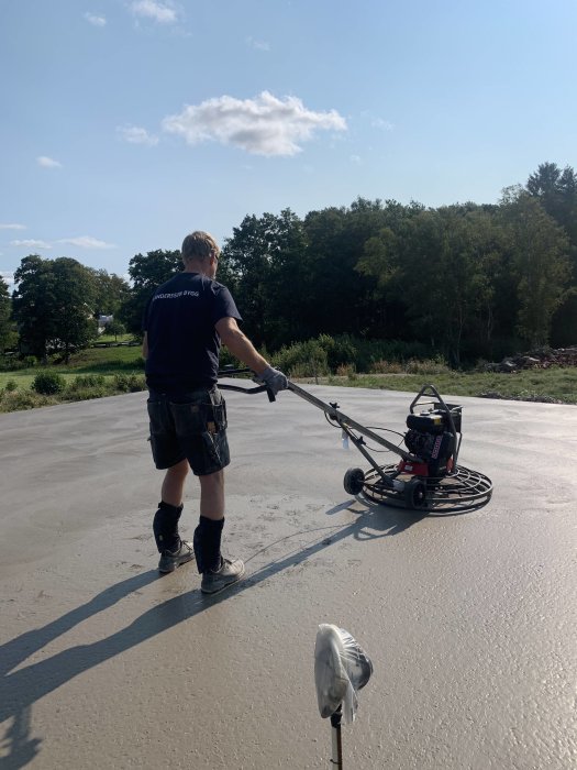 Person som jämnar ut en nygjuten betongyta med en power float under en solig dag.