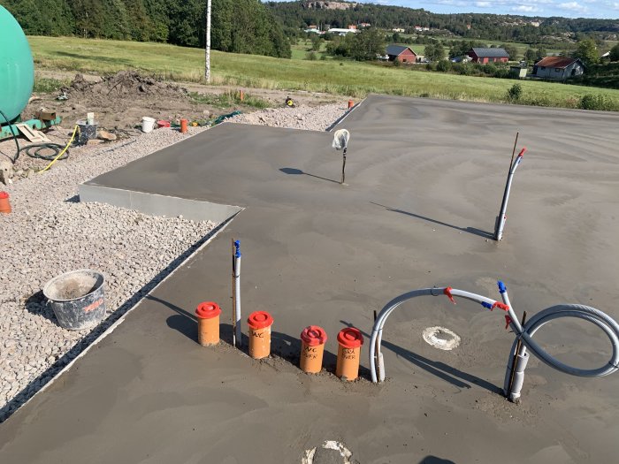 Nylagt betonggolv med rörsystem och avstängningskäppar på en byggplats, omgiven av grus och verktyg.