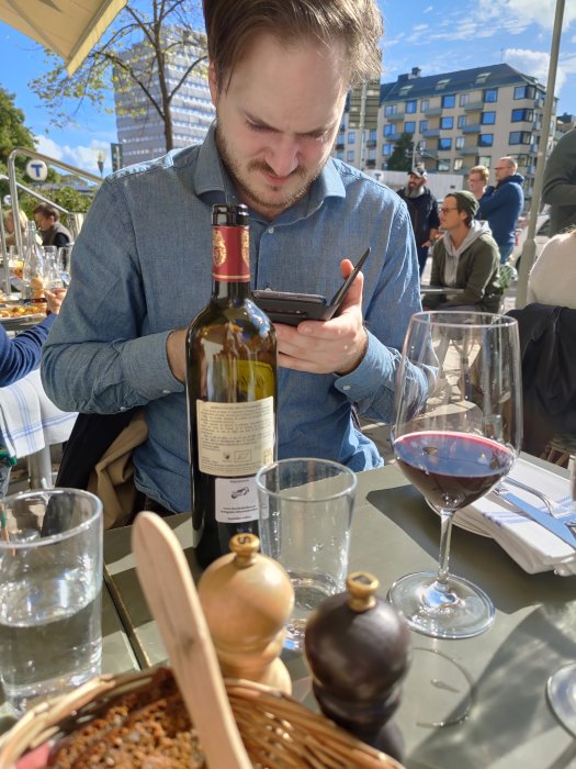 Person använder mobilapp vid lunch med vinflaska och glas på bordet utomhus.