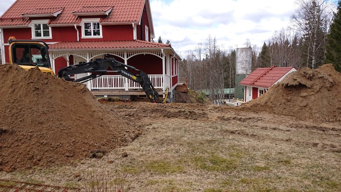 En grävskopa gräver en grop bredvid ett rött hus med veranda för en framtida källarutbyggnad.