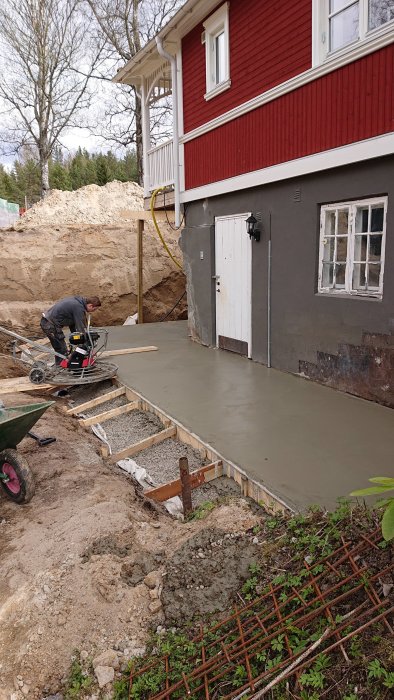 En person jämnar ut nygjuten betong för grunden till en husutbyggnad, med armeringsjärn i förgrunden.