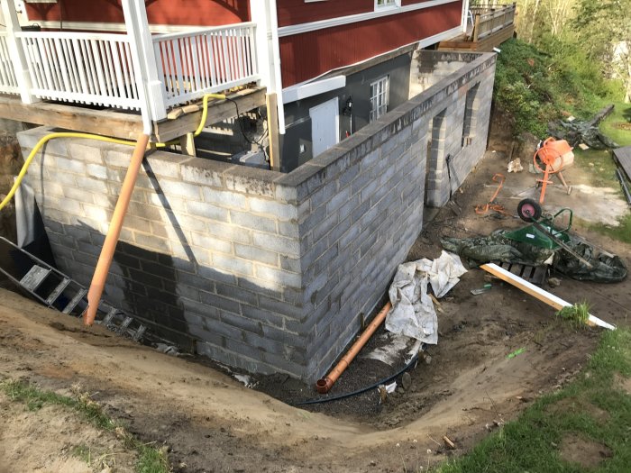 Nybyggd husgrund med tegelmurar, byggmaterial och verktyg framför ett rött hus med veranda.