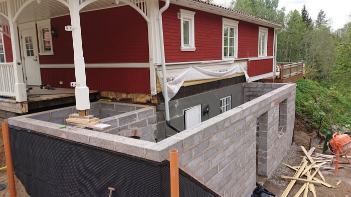 Murningsarbete på en husgrund med oinredd källare intill ett rött hus med vit veranda.