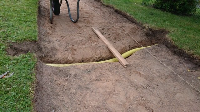 Grävd ränna i trädgård med en gul kabelskyddsrör och en träplanka över rännan.