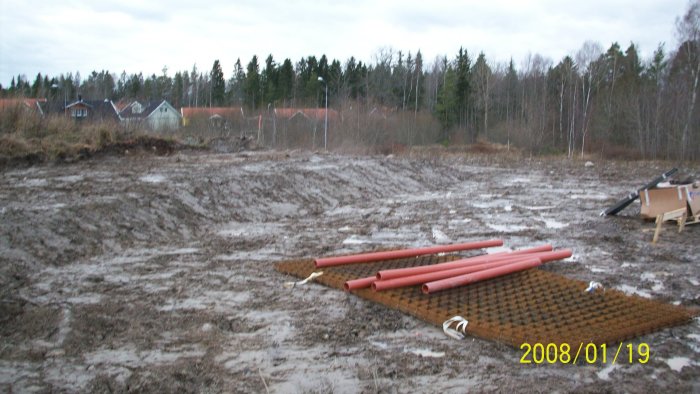 Schaktad och paddad mark med rör och armeringsnät, omgivet av skog och hus.