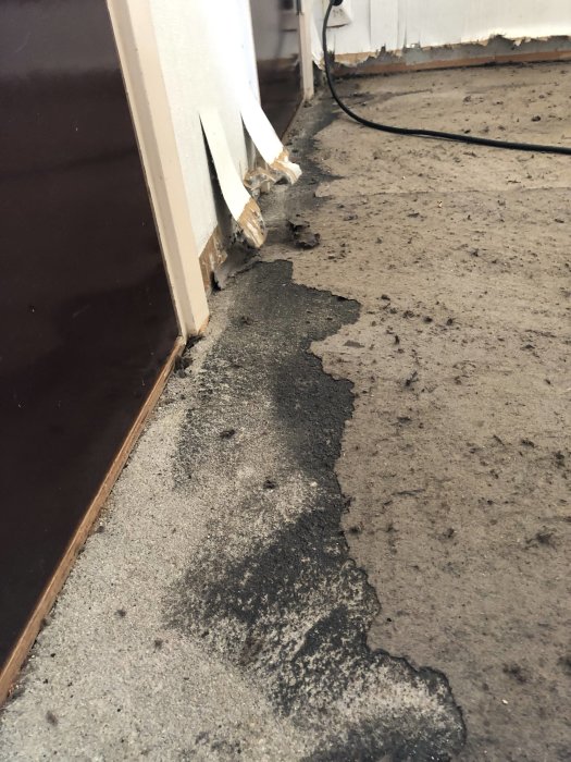 Ett källargolv med rester av borttagen plastmatta och svarta limrester som misstänks innehålla asbest, med strippad papp synlig.