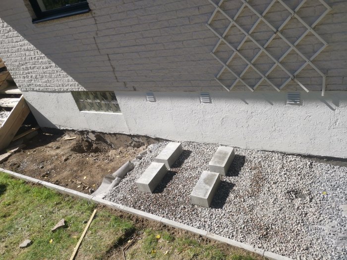 Grävt hål vid husgrund med betongblock, makadam, och installationsrör för pumpen i trädgården.