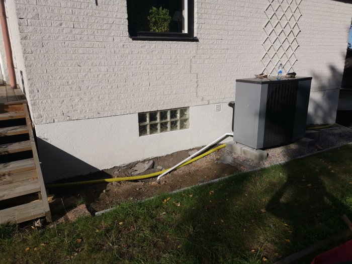 Nyinstallerad pump på betongblock vid husvägg med synliga elkablar och kondensvattenrör, omgivet av grus och grävd jord.