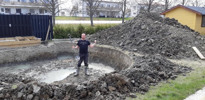 Person står i grävd poolgrop omgiven av högar med utgrävd jord och en spade i handen.