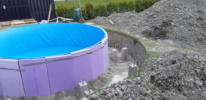 Nygrävd pool med blå presenning och stödbetongfundament, stora högar av grävd blålera i bakgrunden.