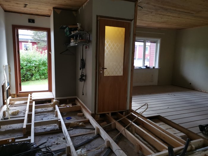 Halvfärdig golvkonstruktion med golvreglar nära en öppen ytterdörr inne i ett hus under renovering, verktyg och byggmaterial synliga.