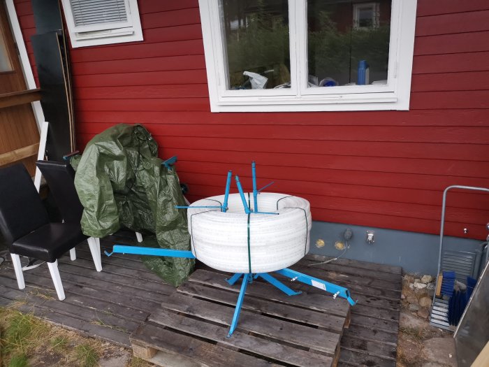 Ett pågående DIY-projekt visas med icke-färdiga vita byggstrukturer stöttade av blåa stödpinnar på en trätrall framför ett rött hus.