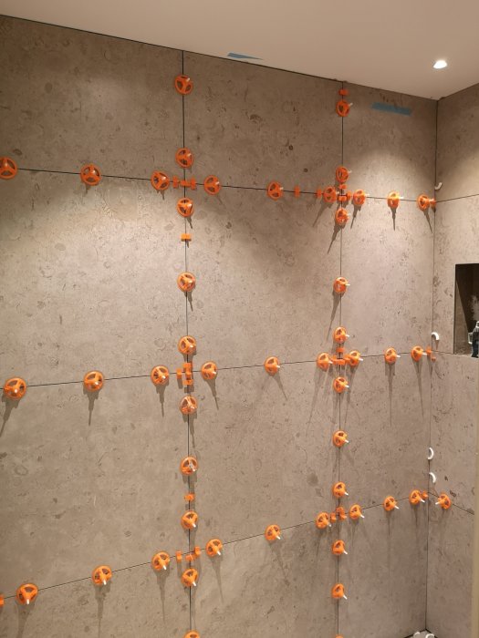 Vägg med beigefärgade kakelplattor i pågående installation, fixerade med orange kryssfogdistanser.