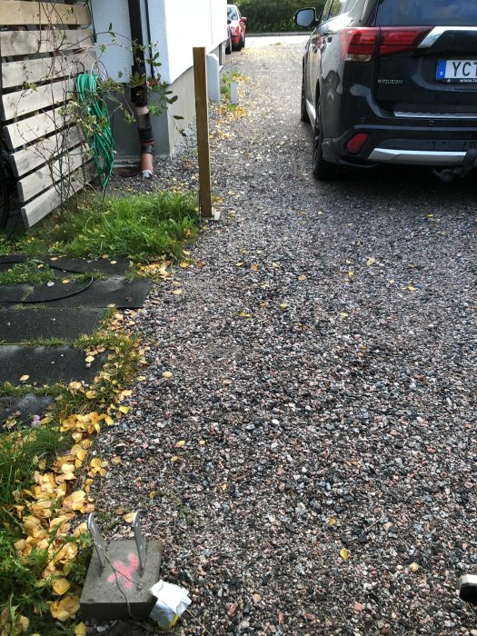 Infart med fyra markerade plintar längs husvägg, avloppsrör synligt, fjärrvärmelåda och parkerad bil.