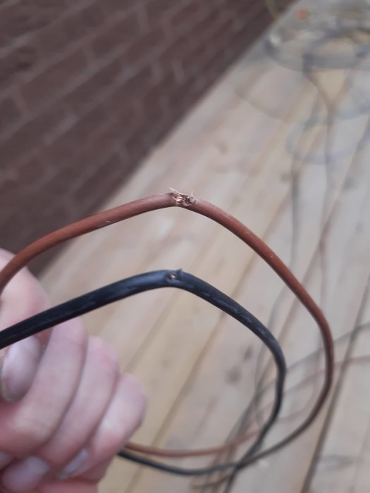 Hand håller en skadad elektrisk kabel med bar koppartråd synlig mot en bakgrund av träaltan och tegelvägg.
