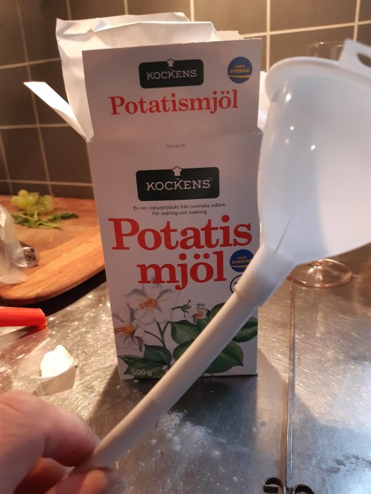 En förpackning Kockens potatismjöl och en vit slev på en köksbänk med skuren grönsak i bakgrunden.