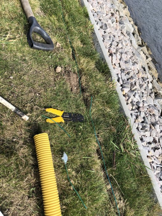 Grävt parti av gräsmatta nära kantsten, med en spade, gult rör och verktyg som ligger utspridda på marken.