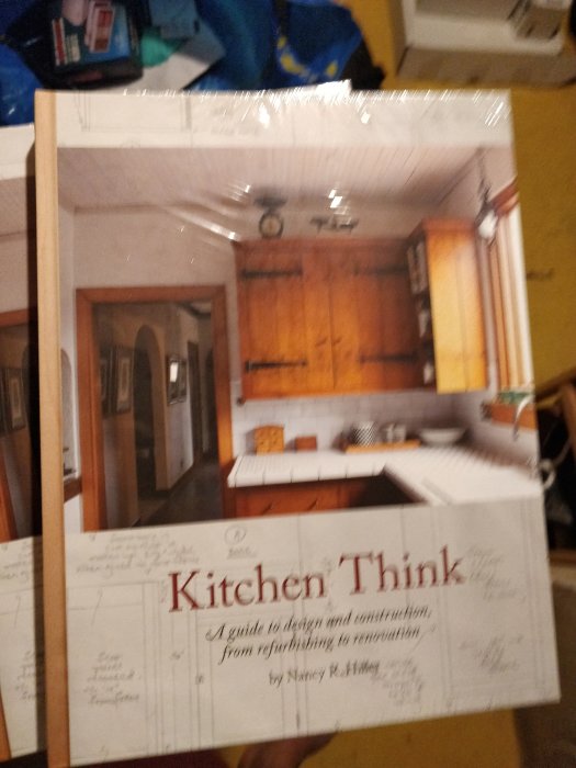 Oskarp bild av en bok med titeln "Kitchen Think" som handlar om köksdesign och renovering, med oskarpa föremål i bakgrunden.