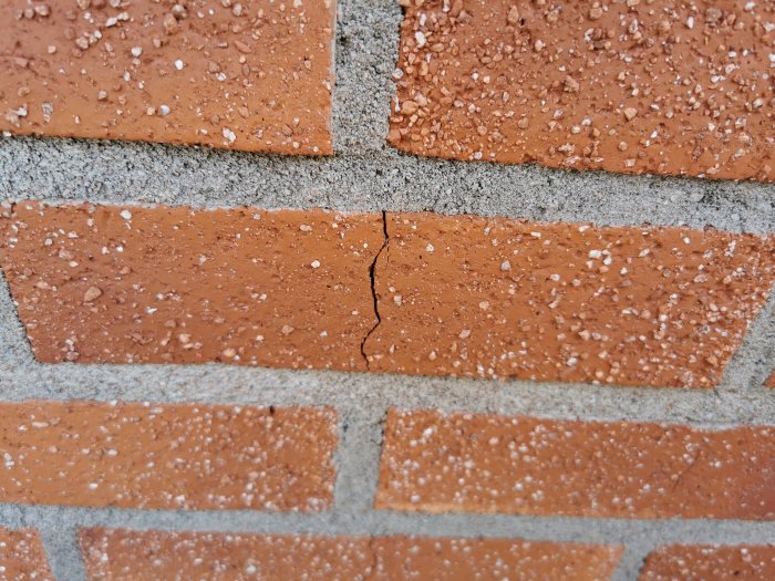 Närbild av en sprucken tegelsten i en vägg, omgiven av intakta tegelstenar och murbruk, möjlig frostsprängning.