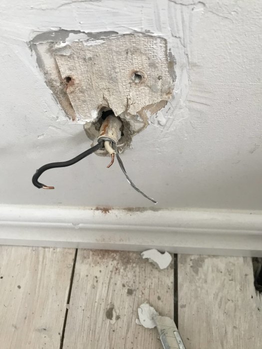 Ett trasigt utanpåliggande vägguttag med svart och vit kabel samt en oskyddad jordtråd framstickande ur en vägg.