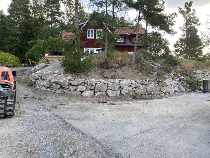 Nybyggd stenmur med sprängsten vid en konstruktionsplats, grävmaskin och hus i bakgrunden.