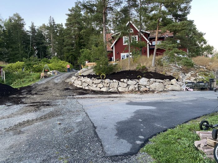 Uppfart med nybyggda stenmurar av sprängsten med en utgrävd yta för pool, röd husfasad i bakgrunden.