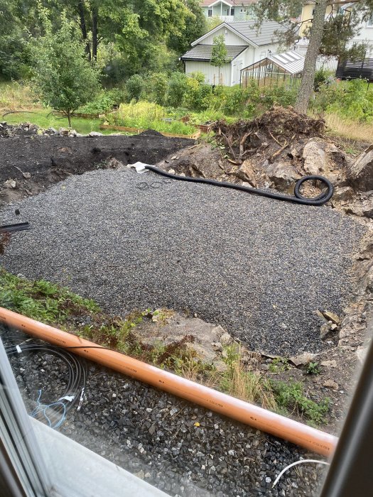 Område i trädgård med sprängsten redo för poolbygge, omgivet av grönska och ett växthus i bakgrunden.