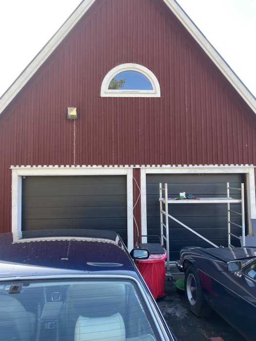 Röd trägaragebyggnad med svart garageport och vit halvmånefönster ovanför, bilar framför och stege vid sidan.