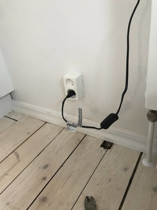 Eluttag i hörn med inkopplad kabel och fungerande installation på vit vägg över ett trägolv.