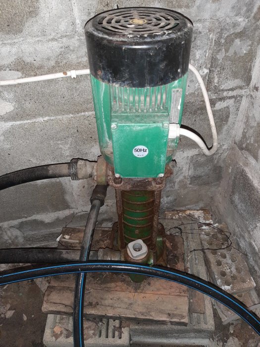 Äldre grön och svart Grundfos CPES 3-80 ejektorpump kopplad till rör och sladdar, placerad i ett stenigt utrymme.