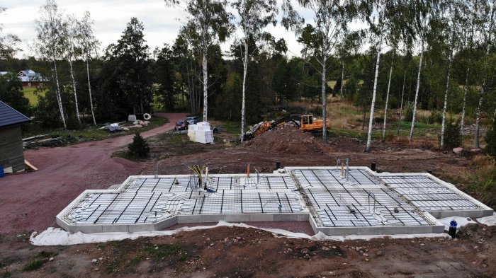 Grund med armering förberedd för gjutning, omgiven av byggmaterial, med skog i bakgrunden.