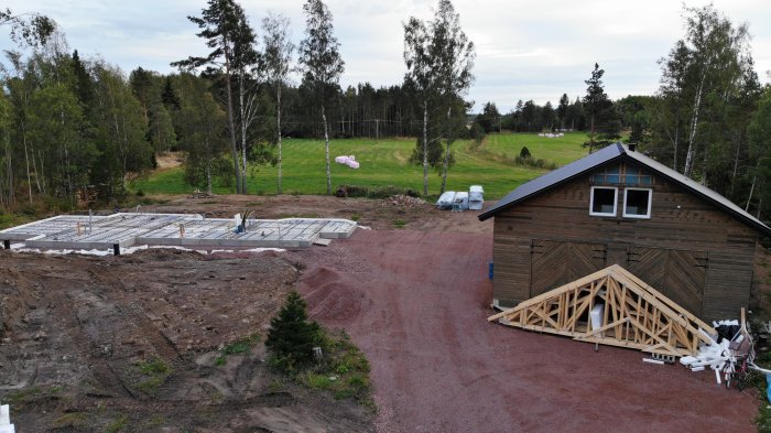 Flygbild över byggplats med förberedd armering för gjutning, takstolar och ett befintligt trähus.