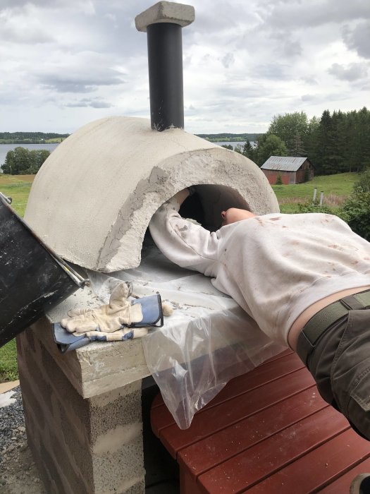 Hemmabyggd pizzaugn med halvfärdig kuppel av eldfast material, utskurna tegel på betongbord och verktyg i en trädgårdsmiljö.