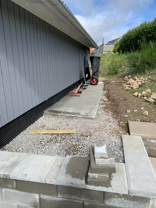 Nybyggd husfasad utan dörrar och skyddsplåtar, byggmaterial och skottkärra i förgrunden.