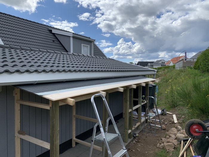 Delvis färdigställt uthus med tak och stötpelare, utan gavelskyddsplåt och dörrar, bredvid en villa.