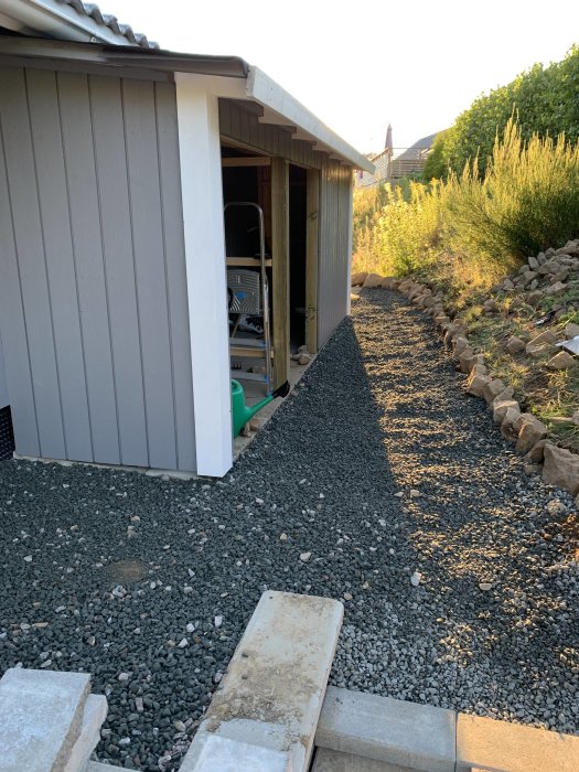 Nybyggd grå uthus utan dörrar, saknar skyddsplåt på gavlarna, omgiven av grusgång och grönska.