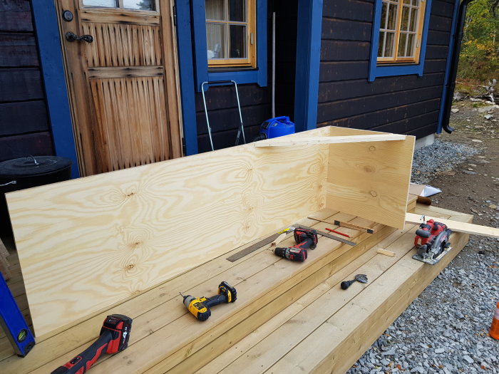 K-plywoodskiva förstärkt med träreglar, verktyg och mätutrustning framför trähus.