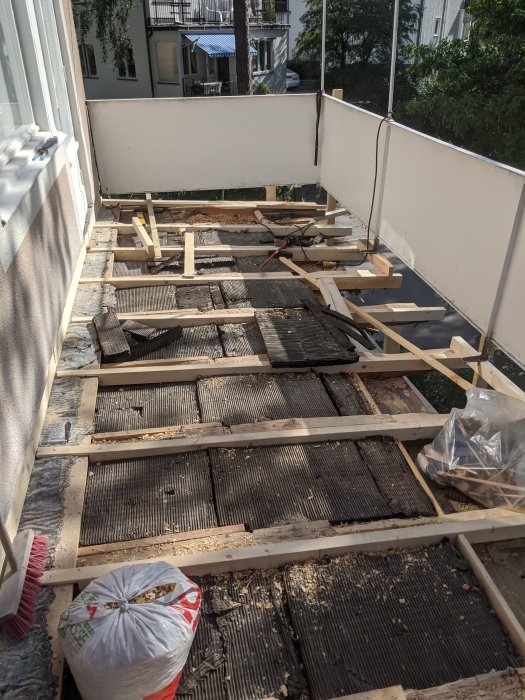 Balkongrenovering i pågående arbete med träreglar installerade och gammalt asfaltlager synligt, i solljus.